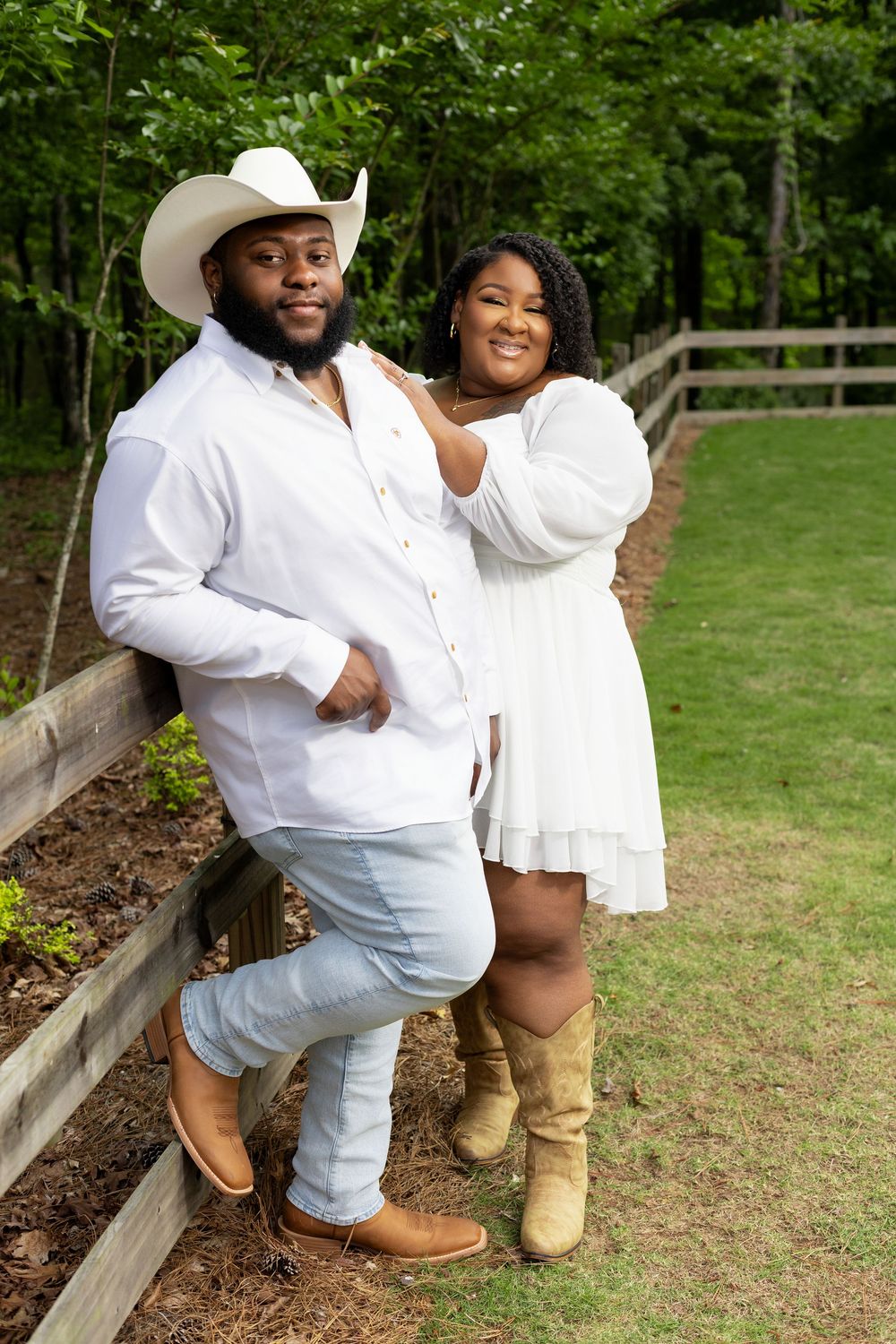 Couple in matching white outfits posing by a wooden fence, man in cowboy hat and woman in boots, outdoors.