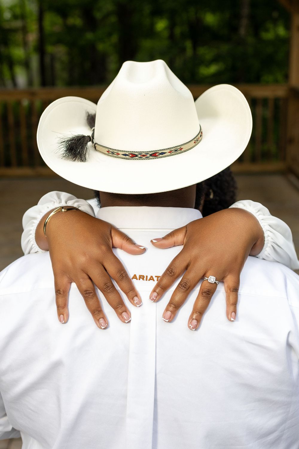 Couple embracing, woman wearing diamond engagement ring hugs man in white Ariat shirt and cowboy hat outdoors.