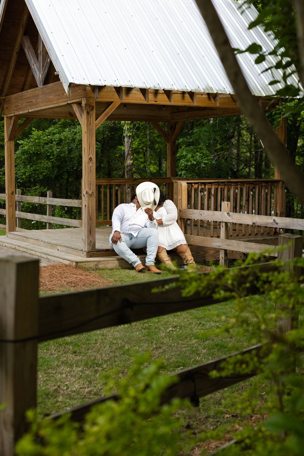 Couple in white outfits sitting together on a rustic wooden gazebo deck surrounded by lush green trees.