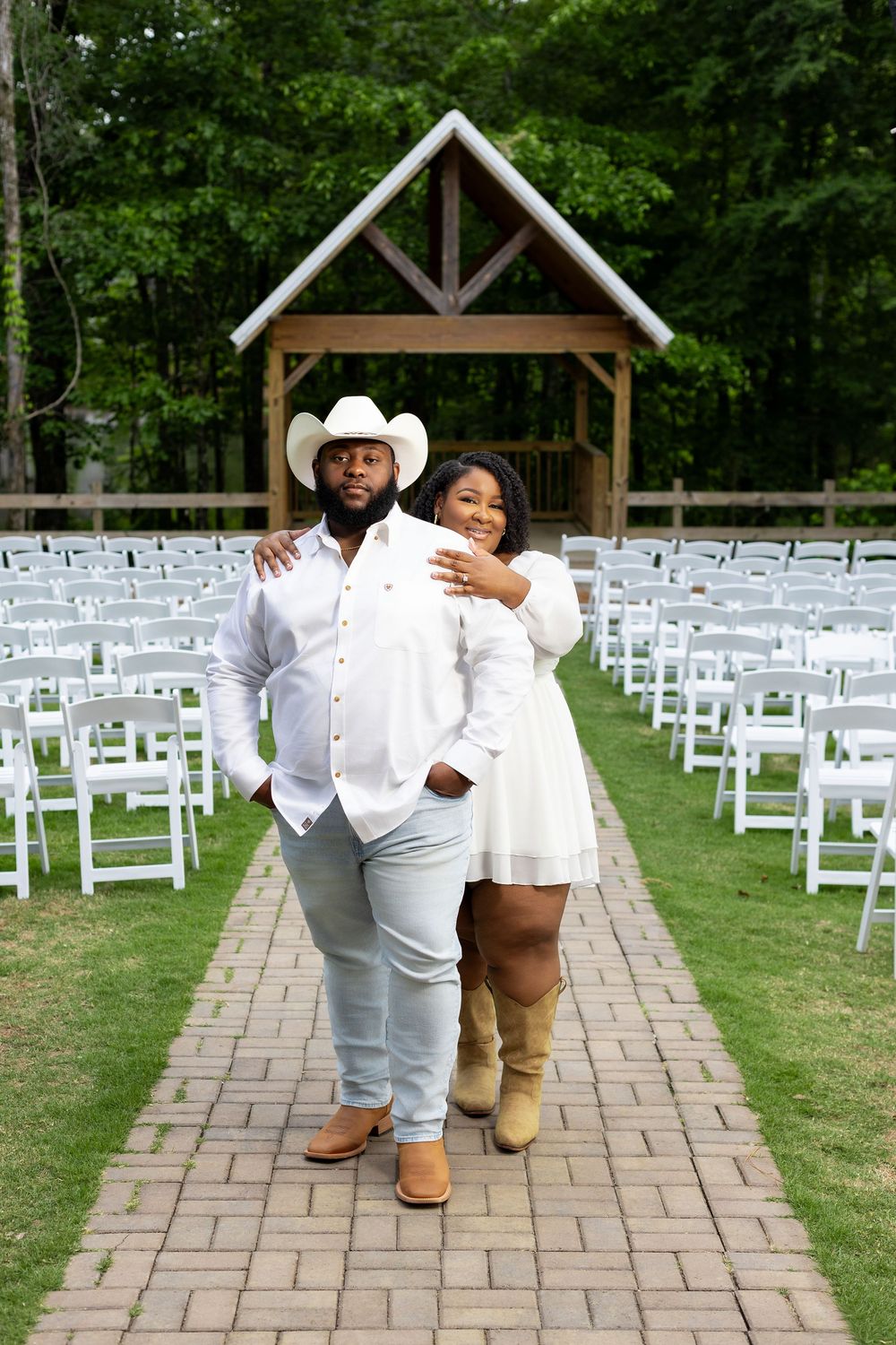 Couple in white western outfits pose at outdoor wedding venue aisle with white chairs and wooden arbor.