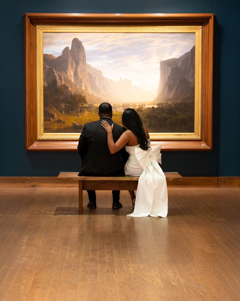 Couple in wedding attire sitting on bench admiring large landscape painting in art museum gallery.