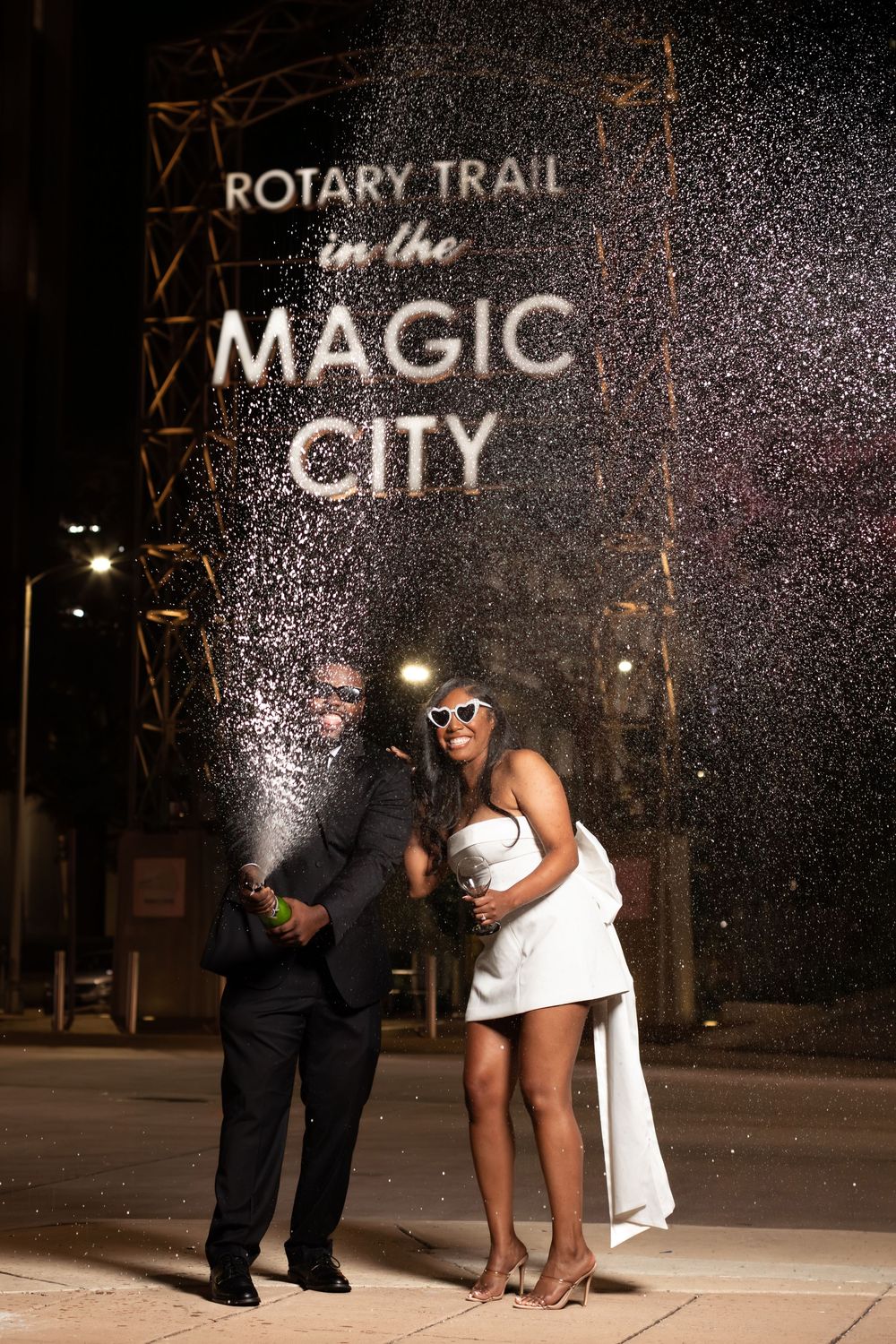 Couple celebrating with champagne spray at Rotary Trail Magic City sign in Birmingham, AL at night.