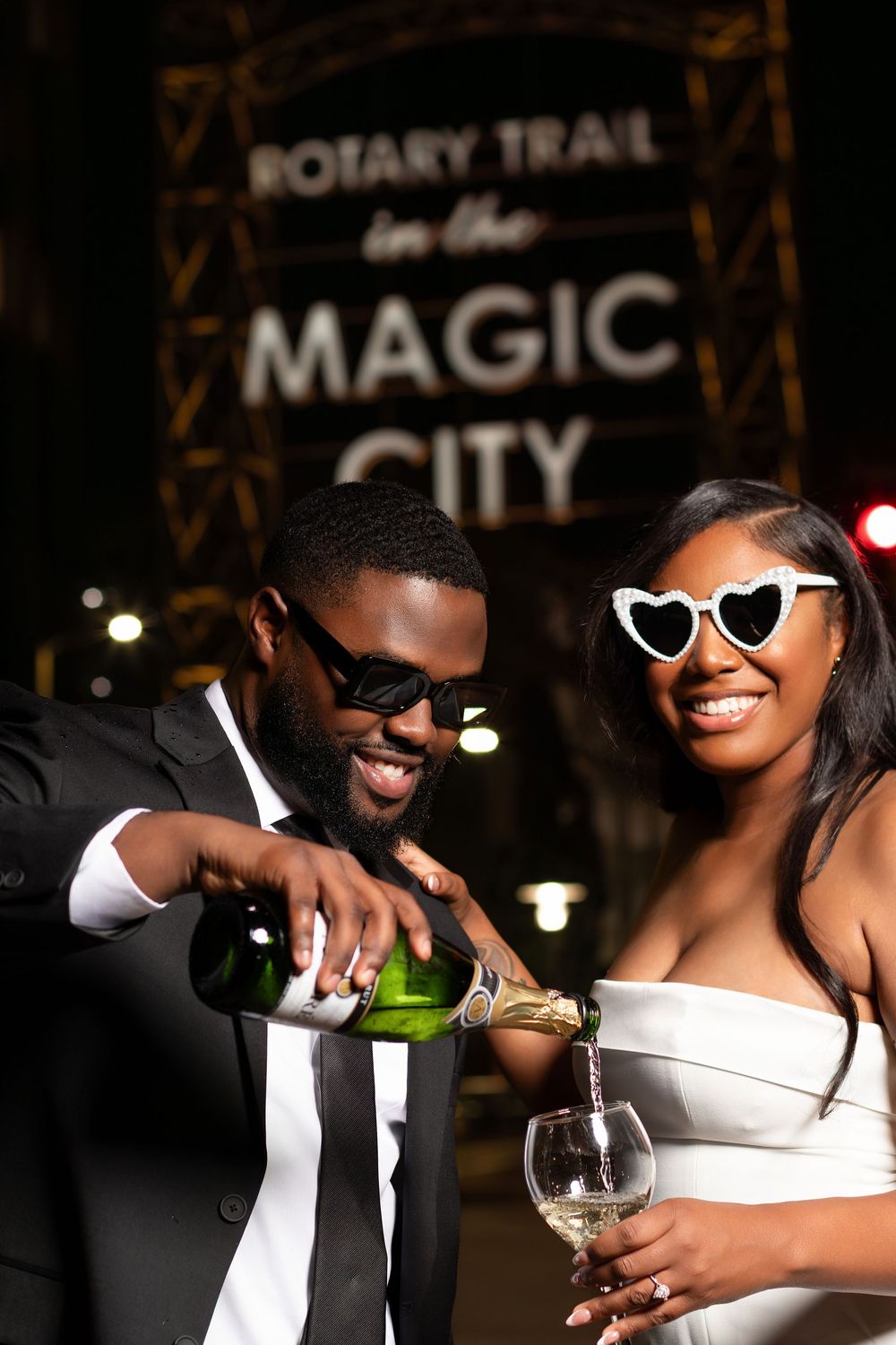 Elegant couple in formal wear celebrating at night near Magic City Rotary Trail sign, pouring champagne.
