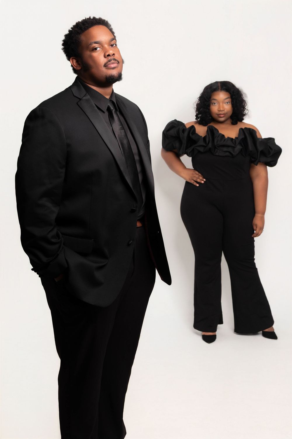 Two people in elegant black formal wear pose against a white background in a professional portrait photo.