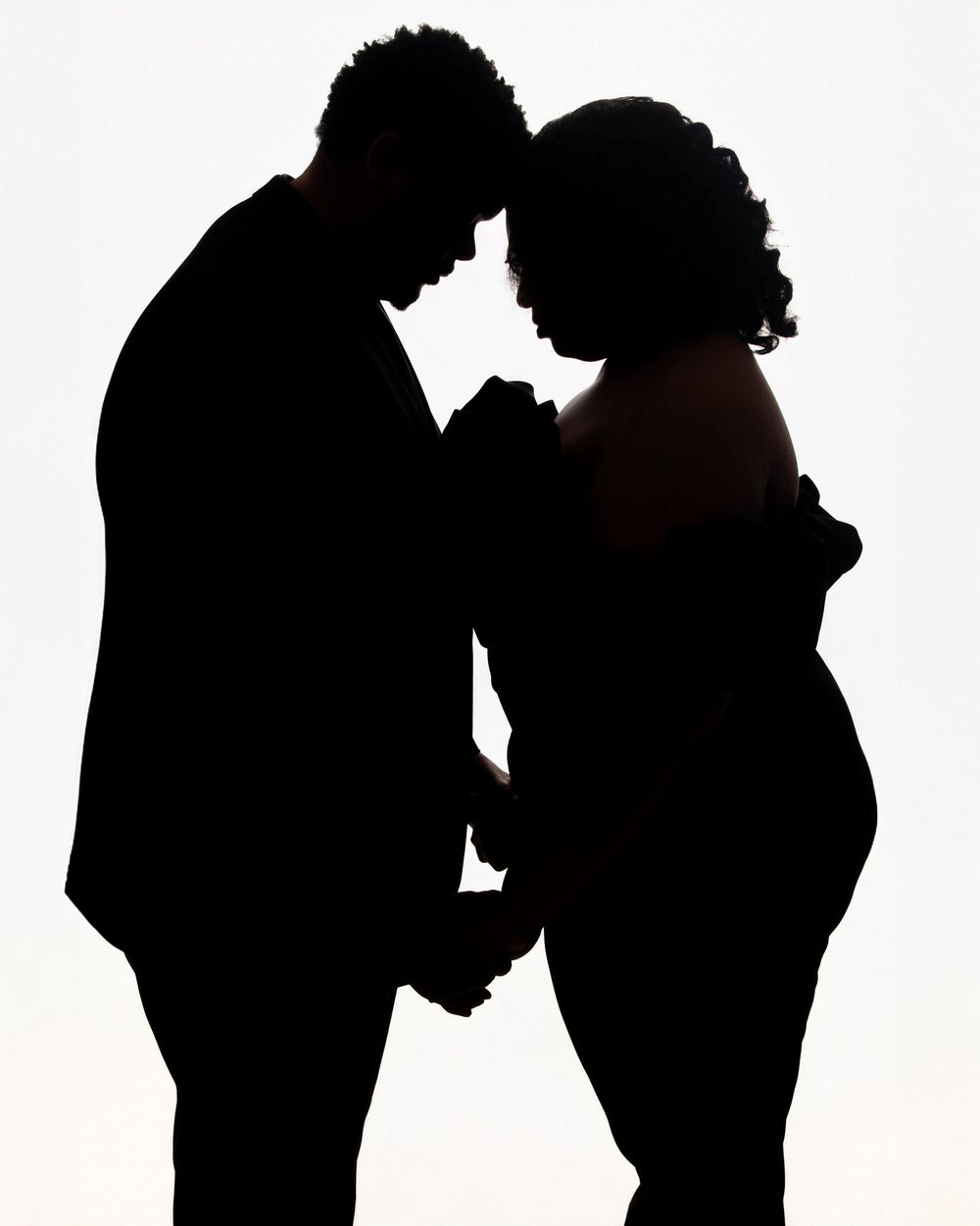 Silhouette of a couple holding hands and touching foreheads, woman with a baby bump, against white background.