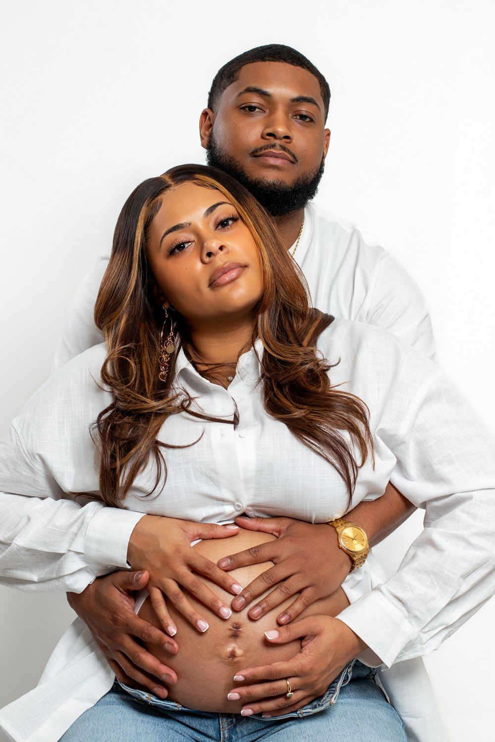 A couple in white shirts poses for a maternity photo, with the man embracing the pregnant woman from behind on white background.