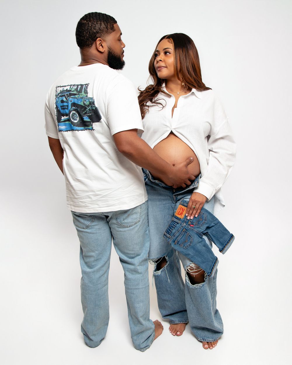 Expecting couple in white shirts and jeans, woman showing baby bump, man touching her stomach tenderly.