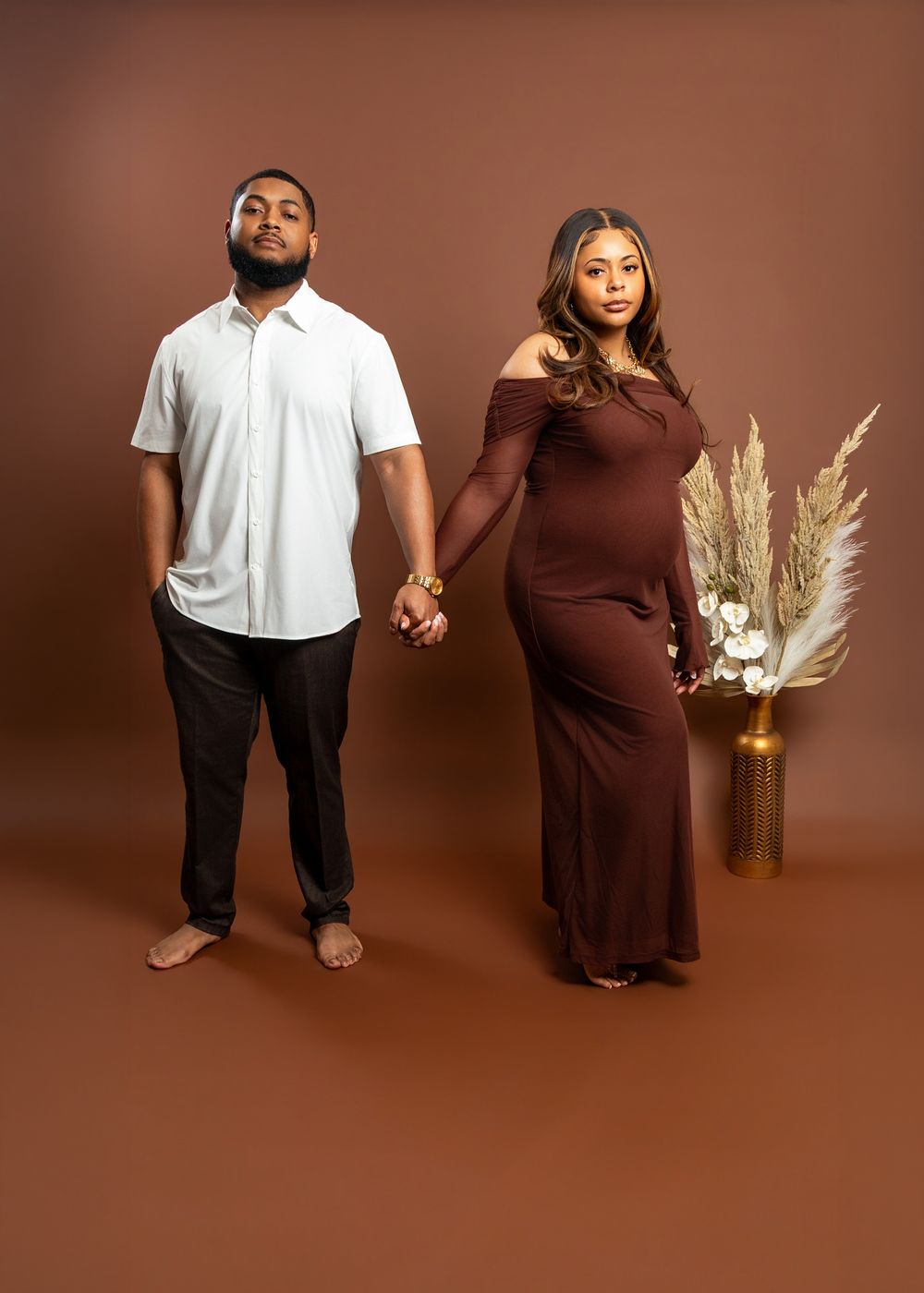 Couple posing for maternity photo shoot, man in white shirt holding hands with pregnant woman in brown off-shoulder gown.