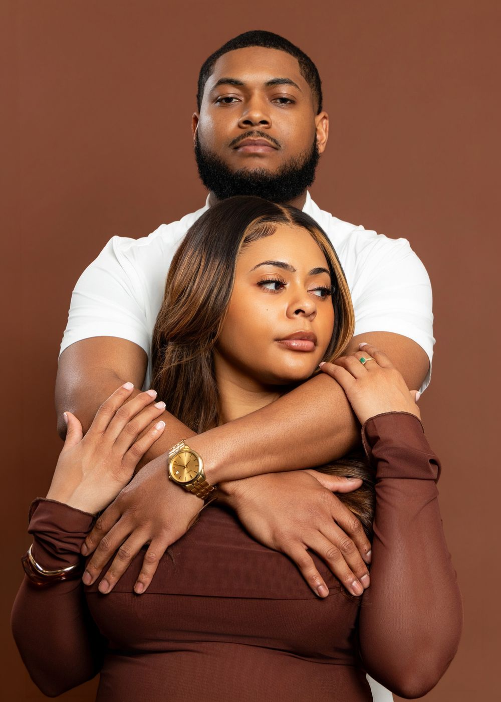 Couple posing together, man embracing woman from behind, wearing brown and white outfits against brown background.