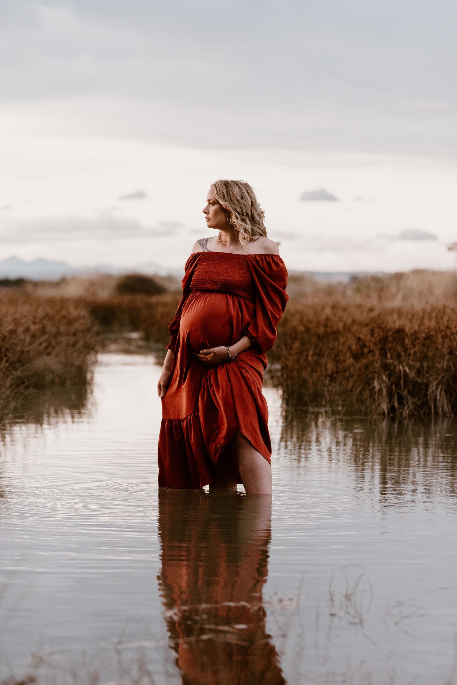 Dramatic maternity portrait in red dress standing in wetland waters during moody sunset.