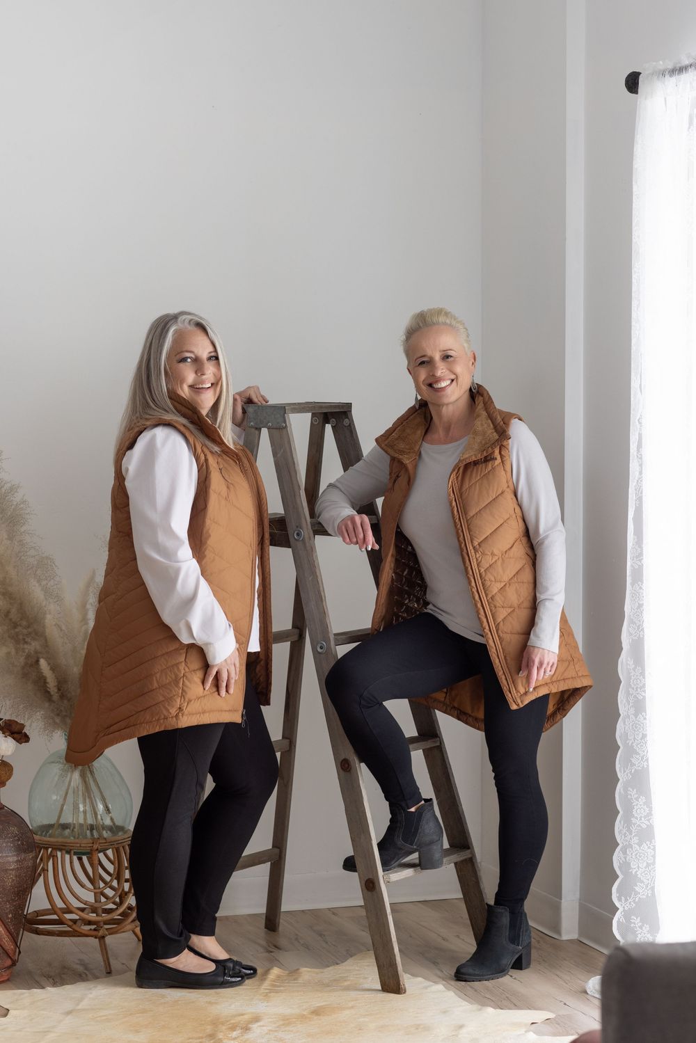 Two women in matching brown quilted vests and white sweaters pose together against a white wall with decorative ladder.