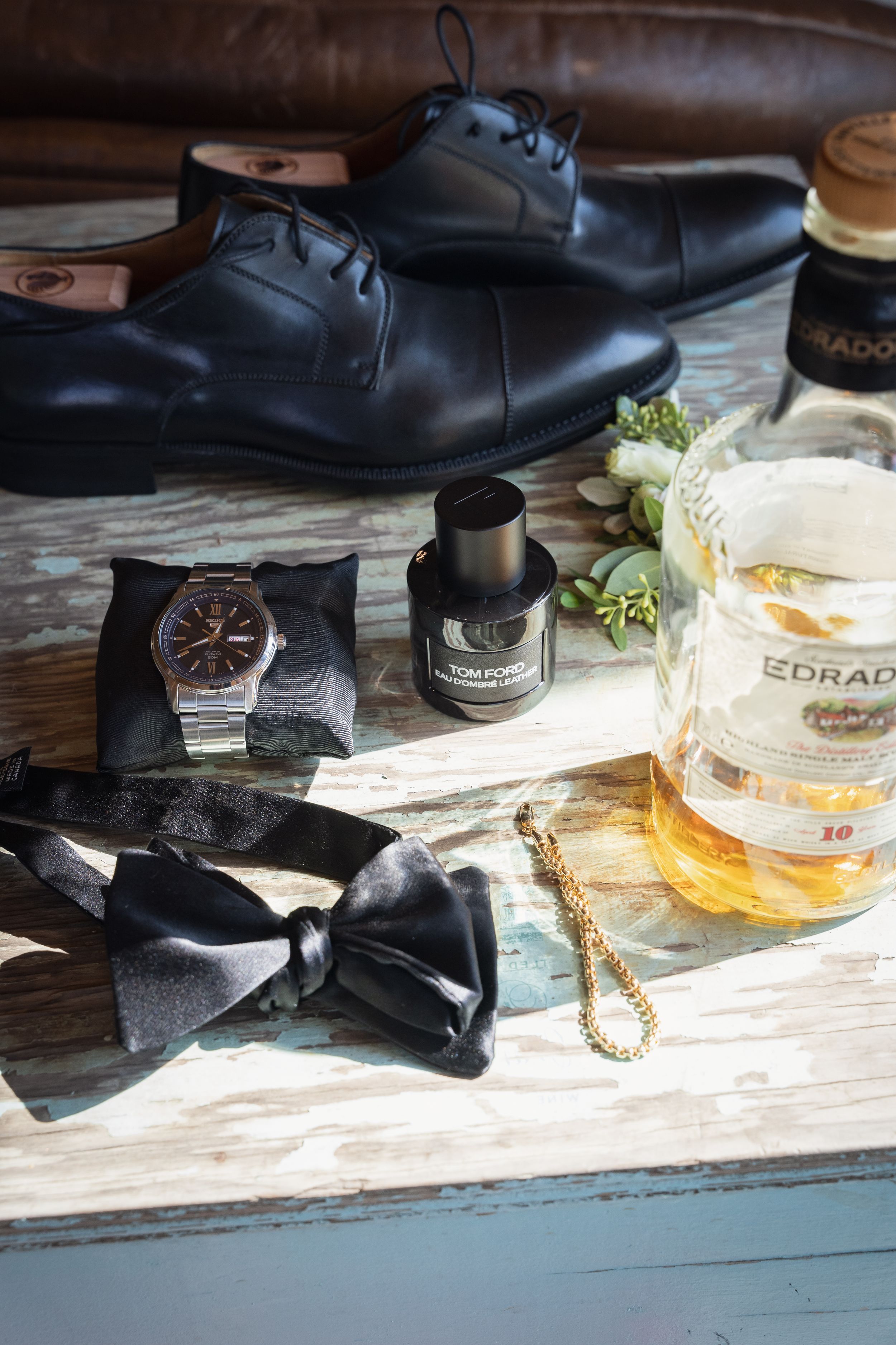 Wedding accessories including dress shoes, bow tie, watch and whiskey on rustic wood.