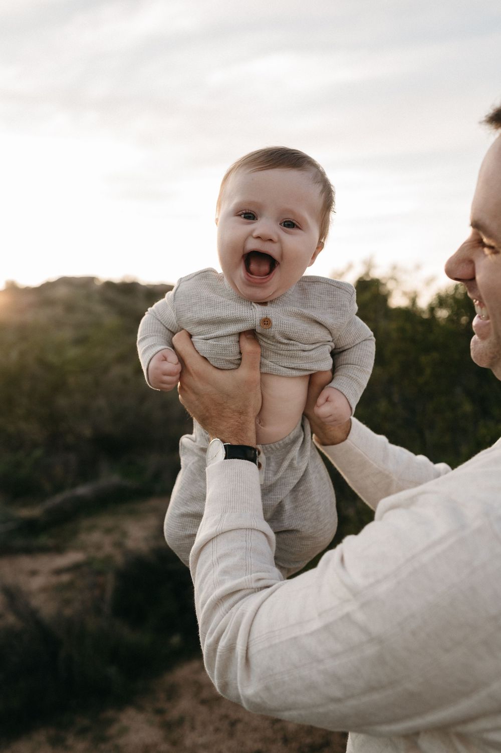 A joyful baby in gray clothing is lifted up by adult hands against a sunset outdoor backdrop.