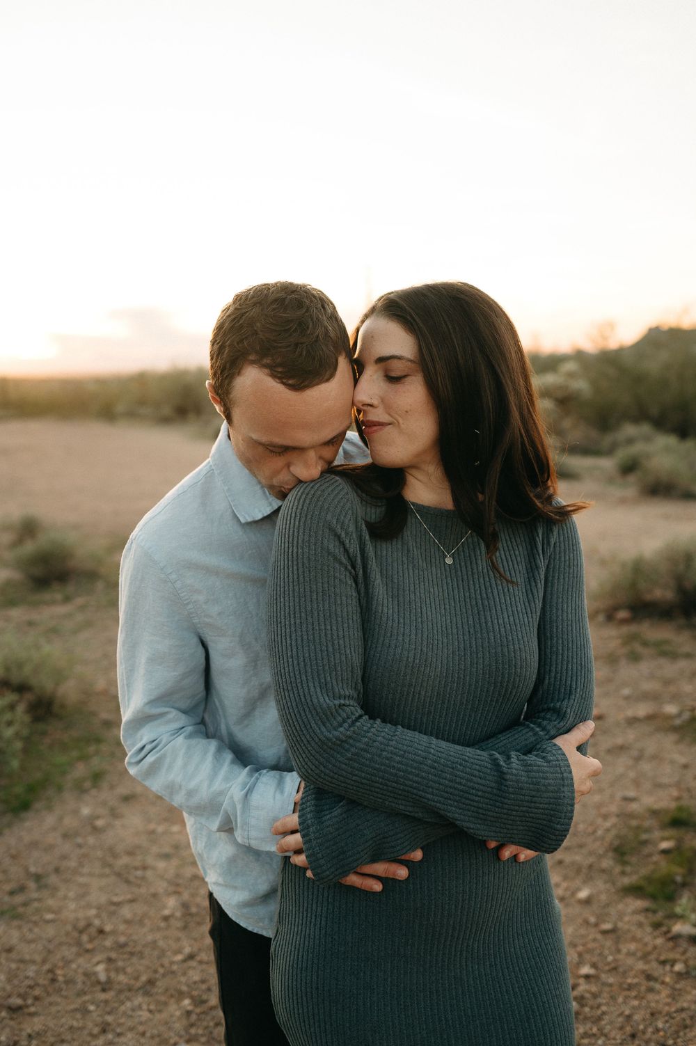 A couple shares an intimate moment at sunset in a desert landscape while wearing casual sweaters.