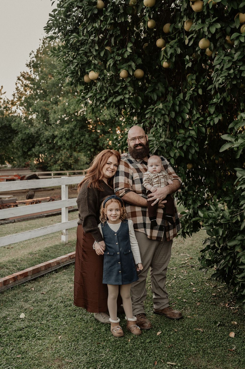 Family enjoying a moment together in an orchard with orange trees and a wooden fence in the background.