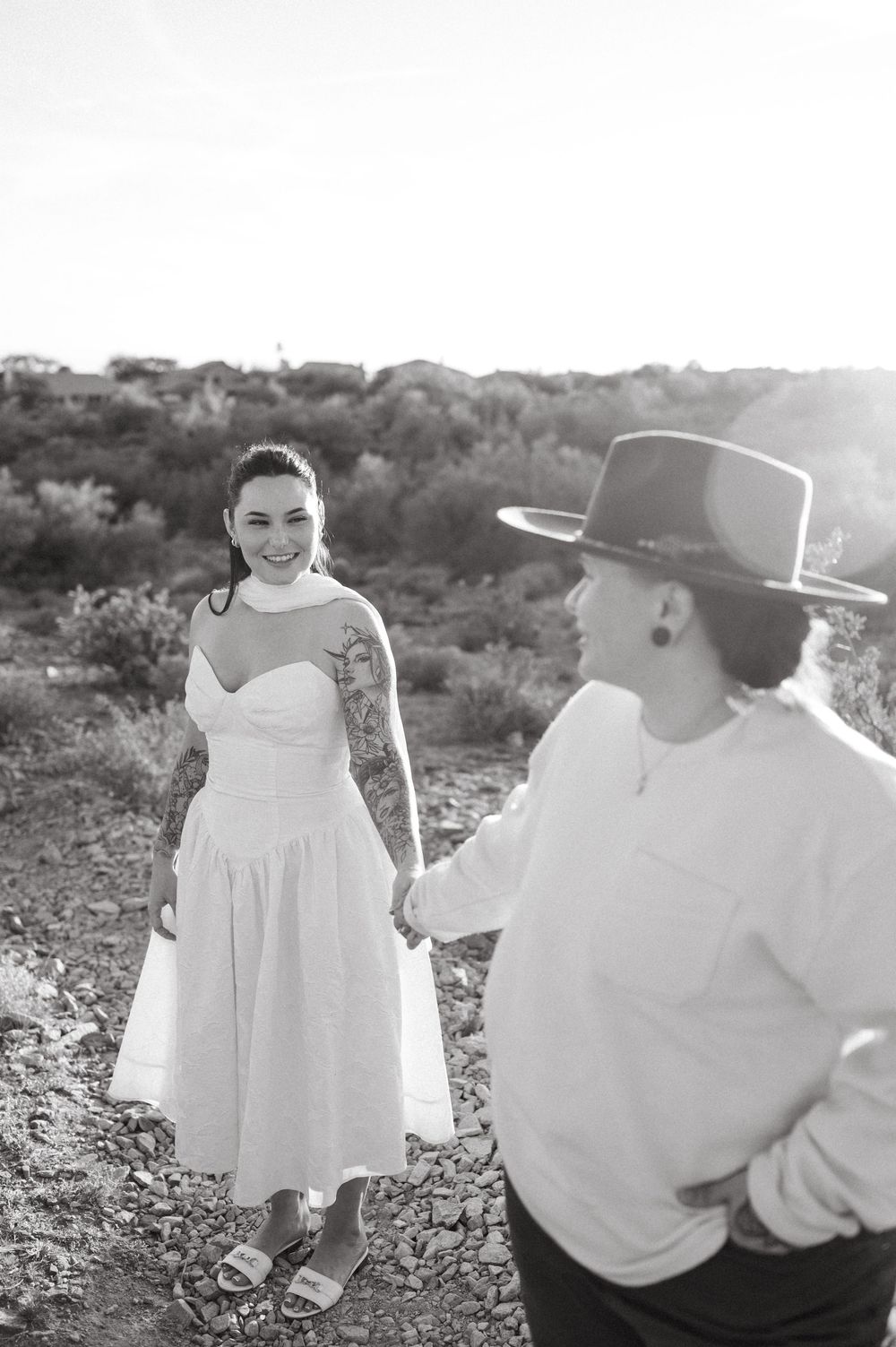 Black and white photo of two brides holding hands in a desert landscape, one in a white dress with tattoos, smiling.