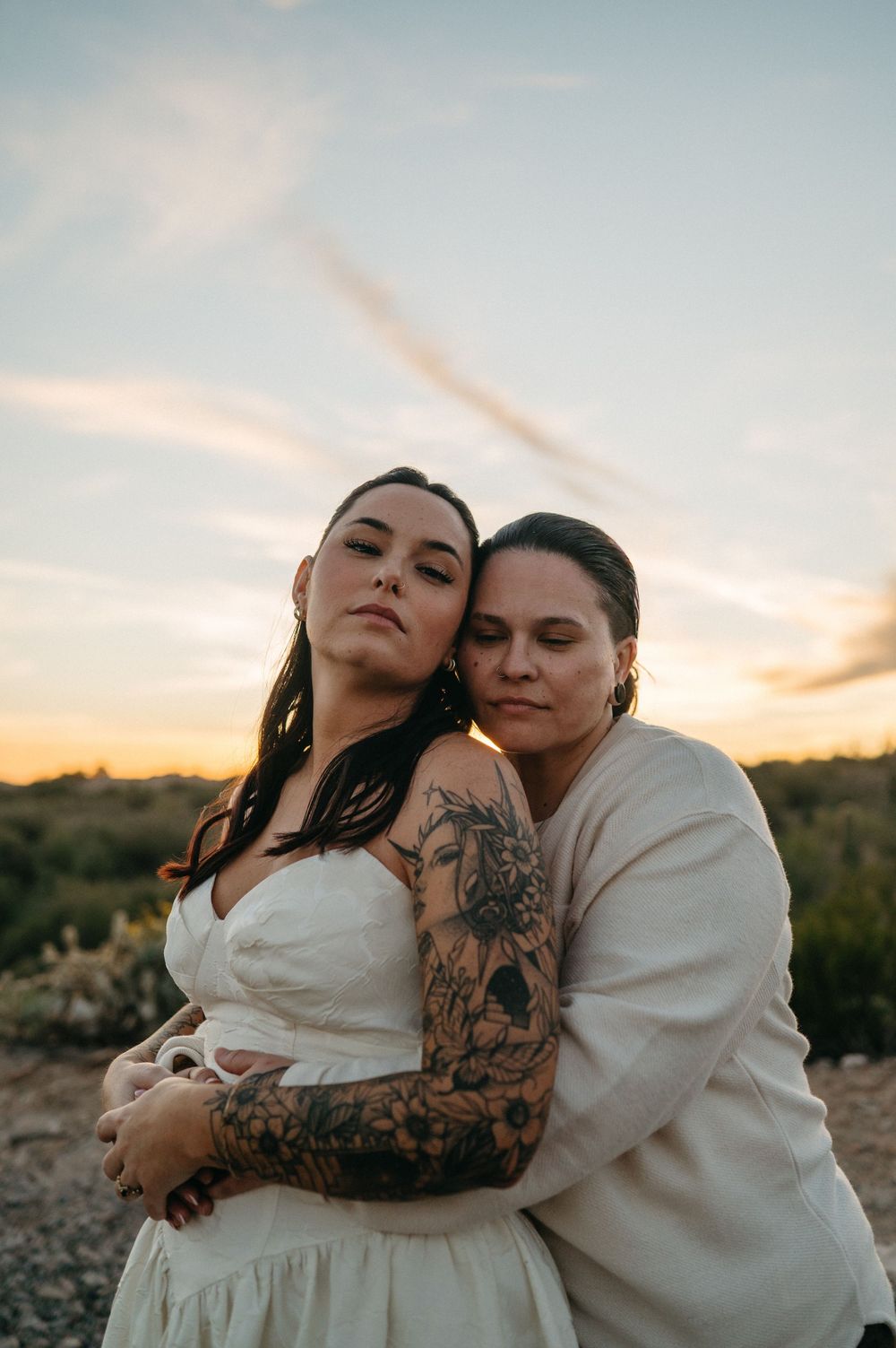 Two women embrace at sunset, one in a white dress with tattoos, sharing an intimate moment outdoors.
