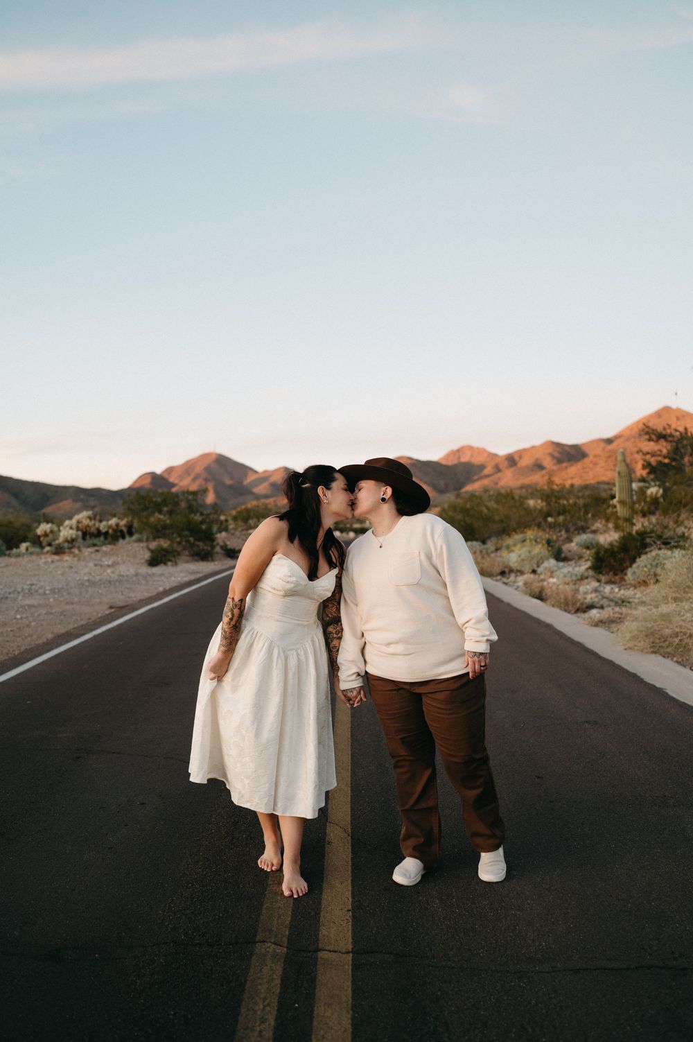 Couple sharing a kiss on a desert road in white dress and cowboy hat with mountains at golden hour.