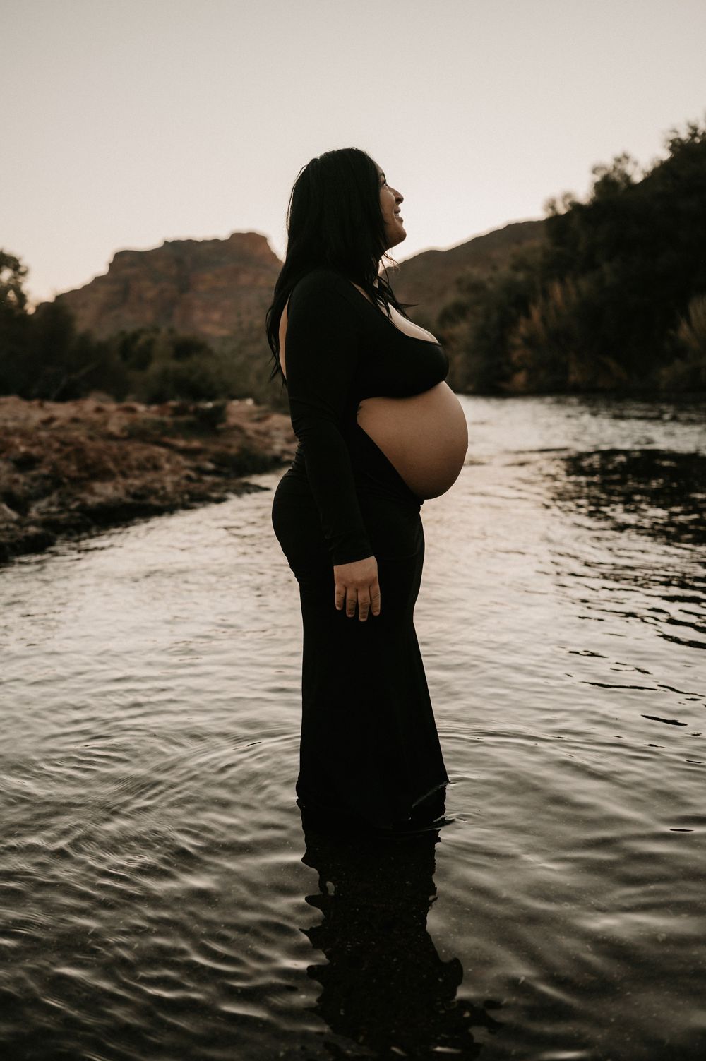 A silhouetted pregnant woman in a black dress stands in shallow water at sunset with rocky cliffs in the background.