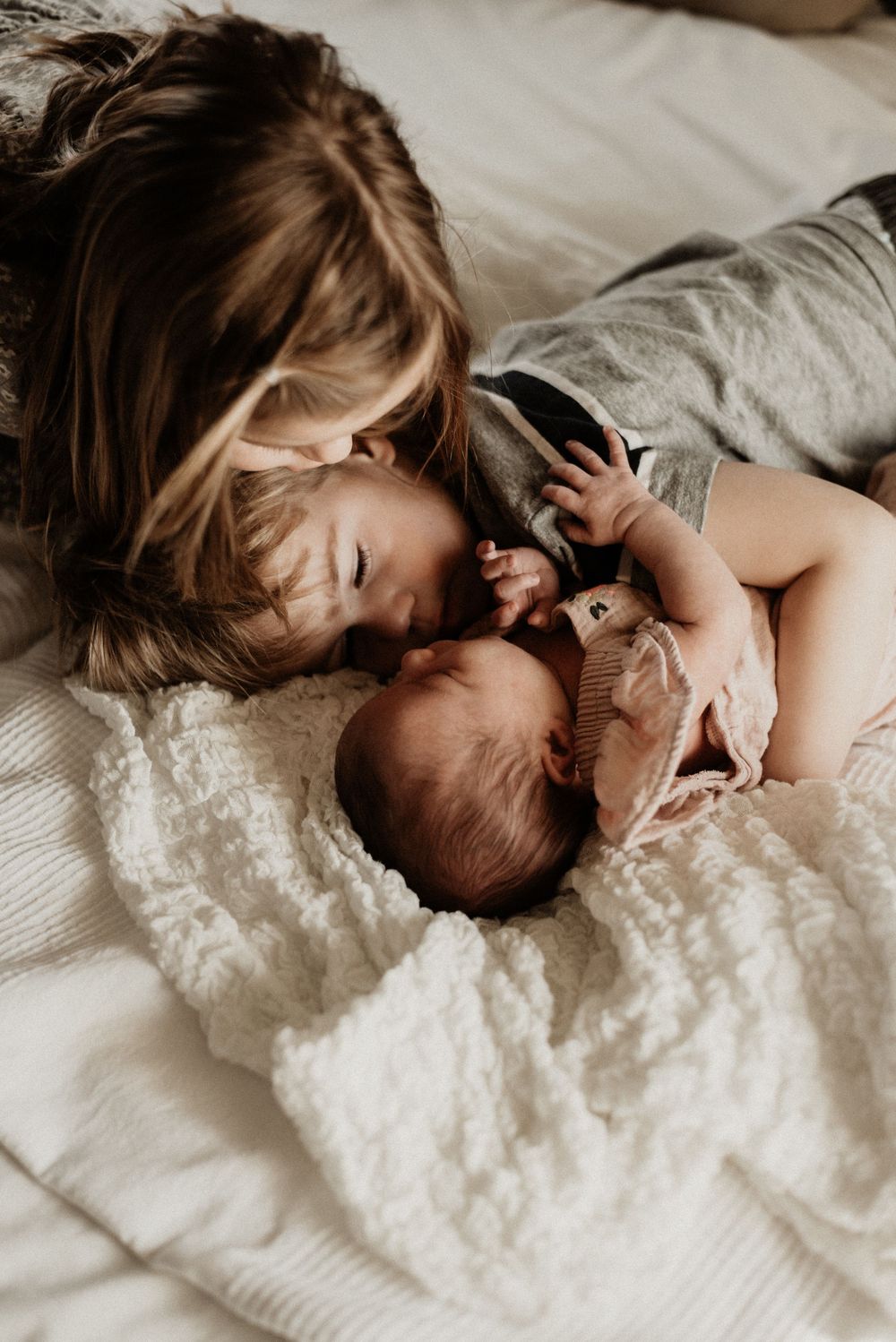 Tender sequence showing siblings bonding with newborn baby on white bedding.