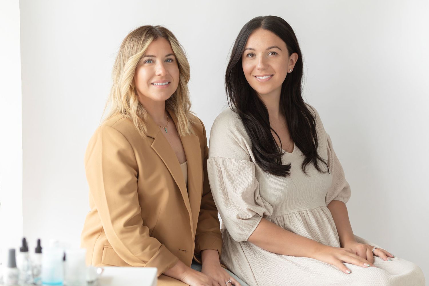 Two women in business casual attire sit together against white background.