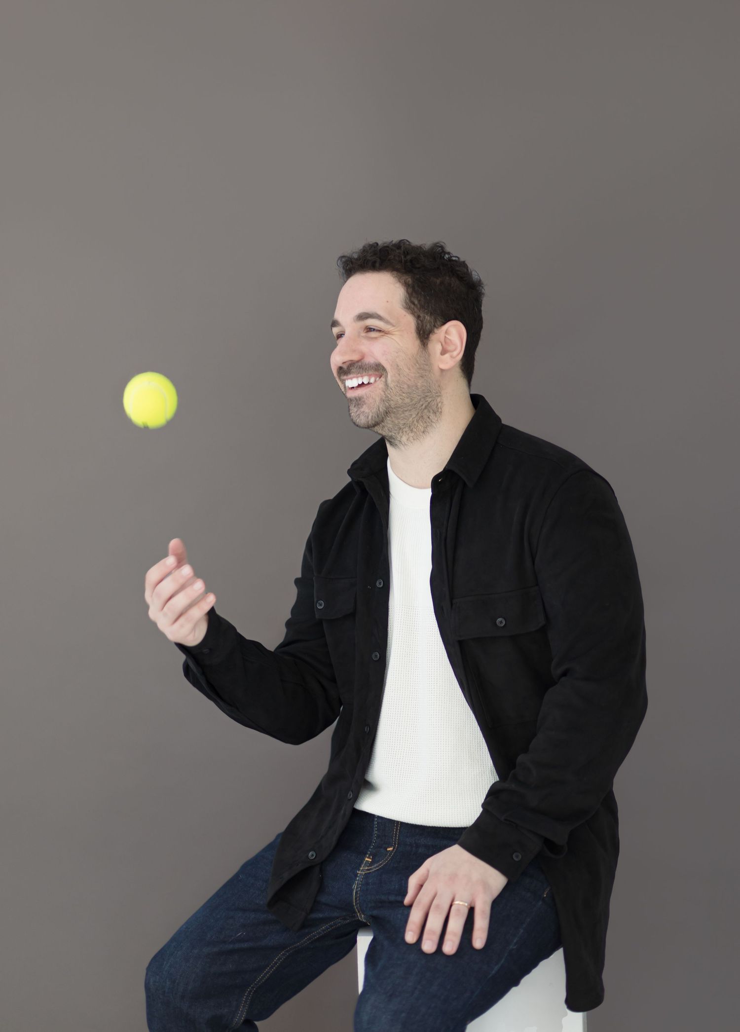 Person tossing a yellow tennis ball in the air while seated and smiling against a grey background.