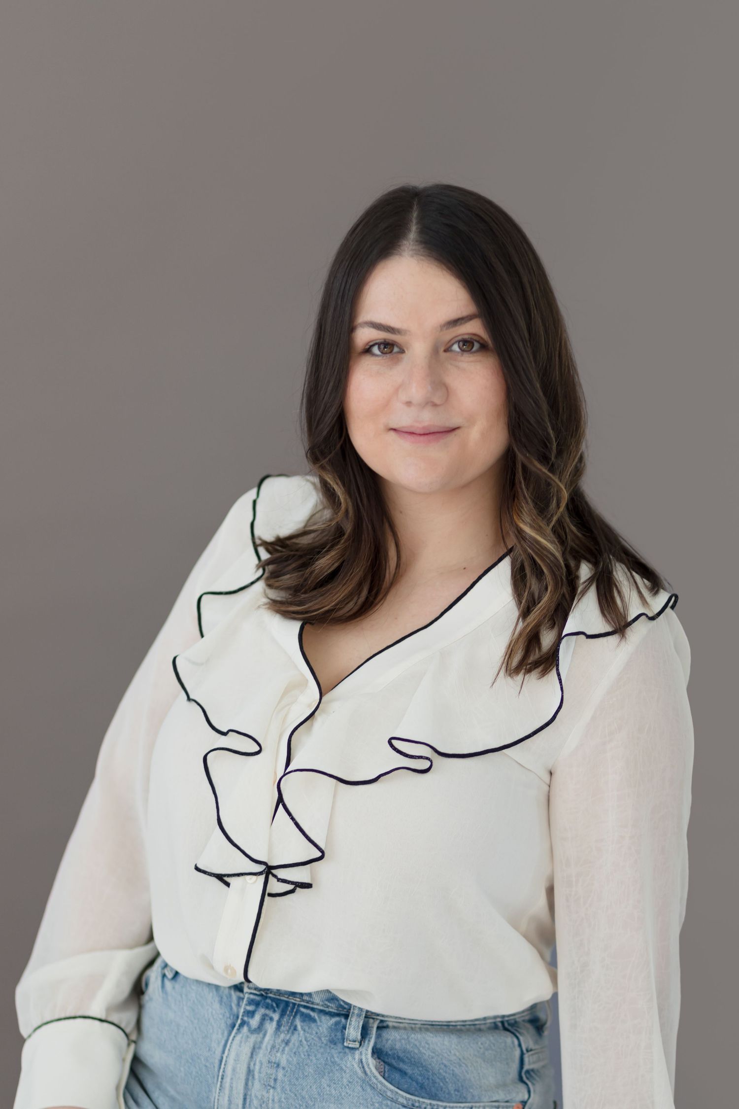 Woman in white ruffled blouse and jeans poses against grey background.