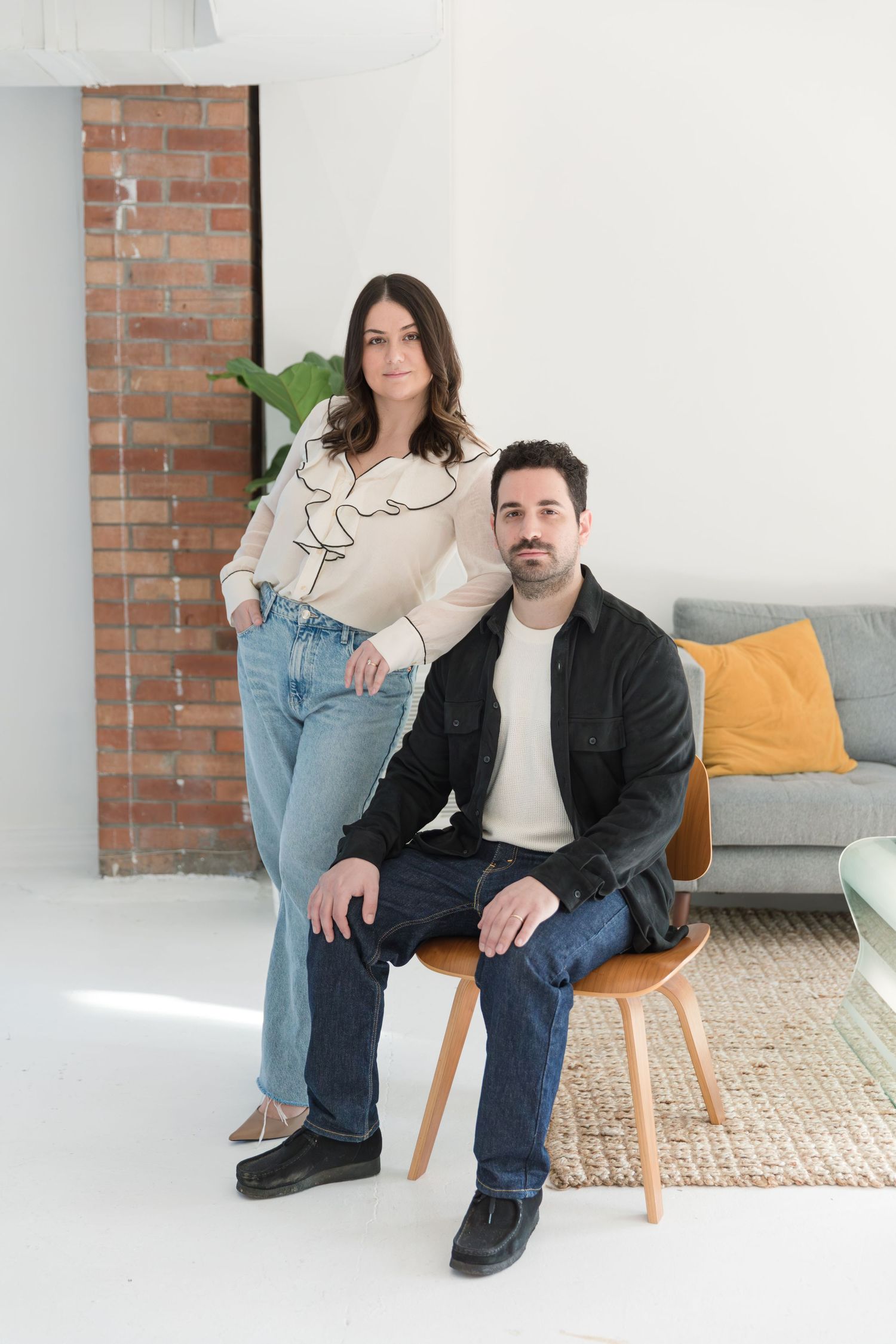 Two people in casual attire sitting in a modern living room with exposed brick and yellow throw pillow.