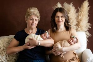 Two people holding newborn babies pose together on a couch with decorative pampas grass in the background.