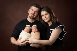 Professional newborn photography series showing tender moments with parents holding sleeping twins in dark tones.