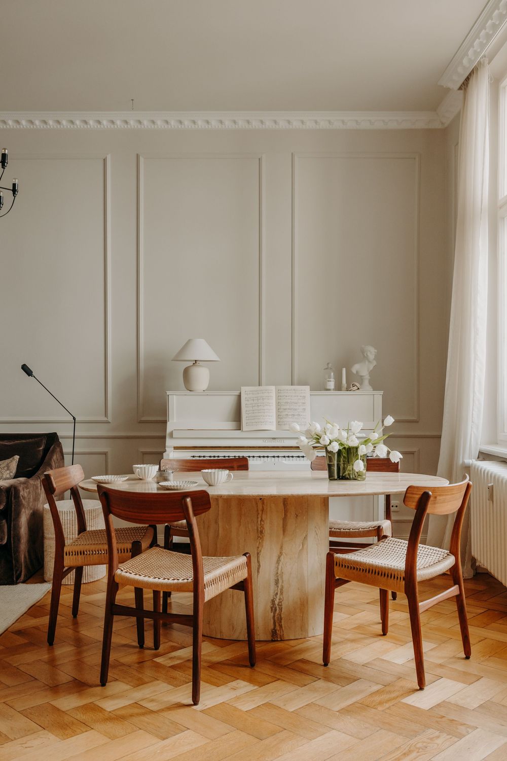 Elegant dining room with round marble table, wooden chairs, white piano, and neutral wall paneling on parquet floors.