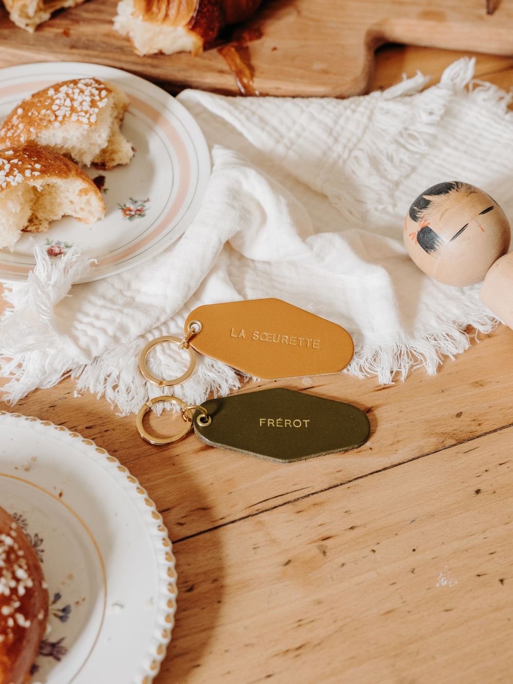 Two gold keychains labeled LA SOUBRETTE and FREROT on a wooden table with pastries and a boiled egg.