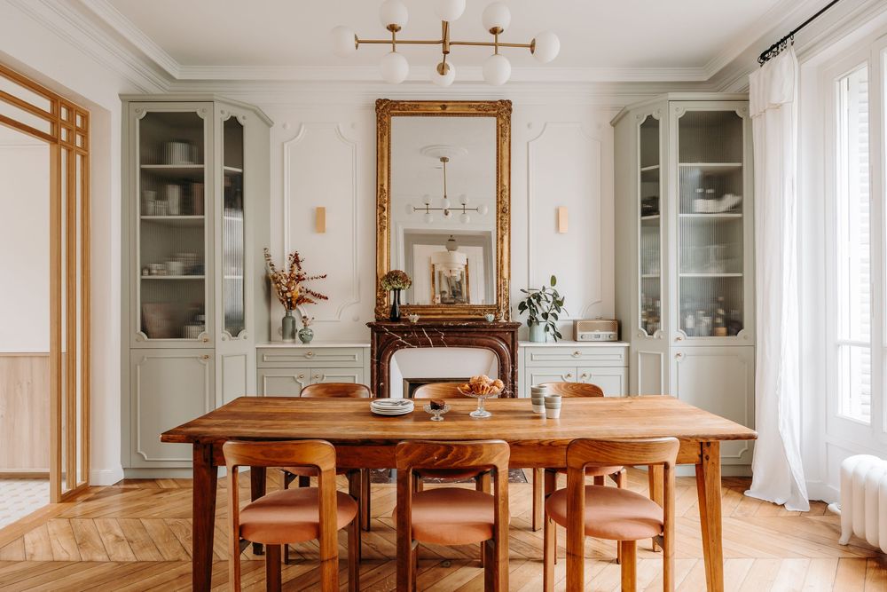 Elegant French dining room with wooden table, sage green cabinets, gold mirror, herringbone floors, and white molded walls.