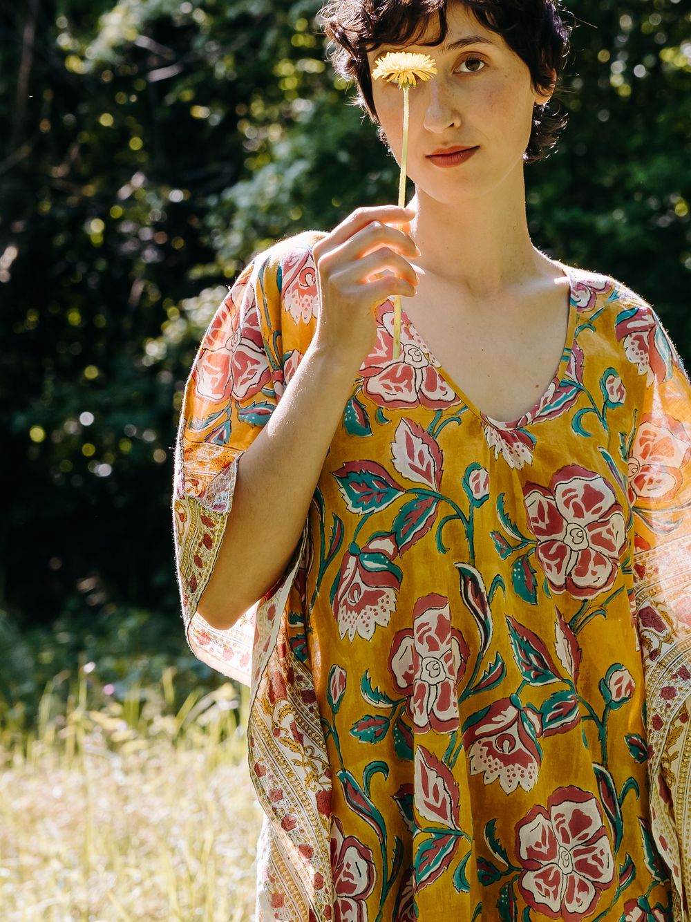Woman in yellow floral maxi dress holding a wildflower outdoors in a sunlit garden setting.
