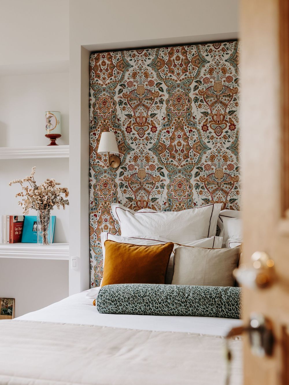 Cozy bedroom nook with ornate floral wallpaper, layered neutral pillows, mustard velvet cushion, and built-in white shelves.