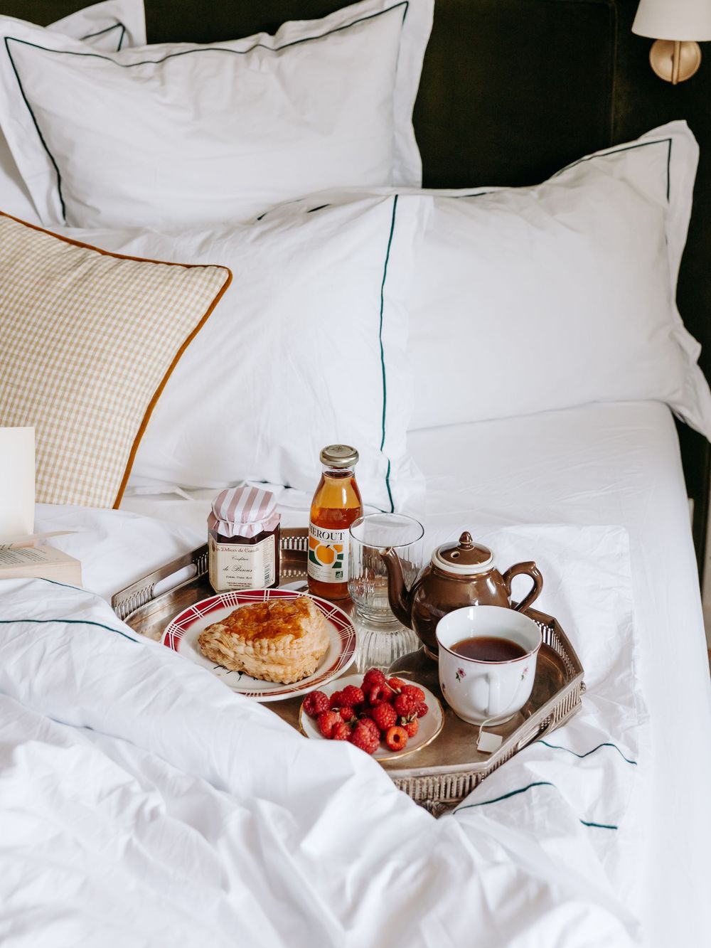 A cozy breakfast tray with tea, pastries, raspberries, jam, and honey served on a white bed with crisp linens.