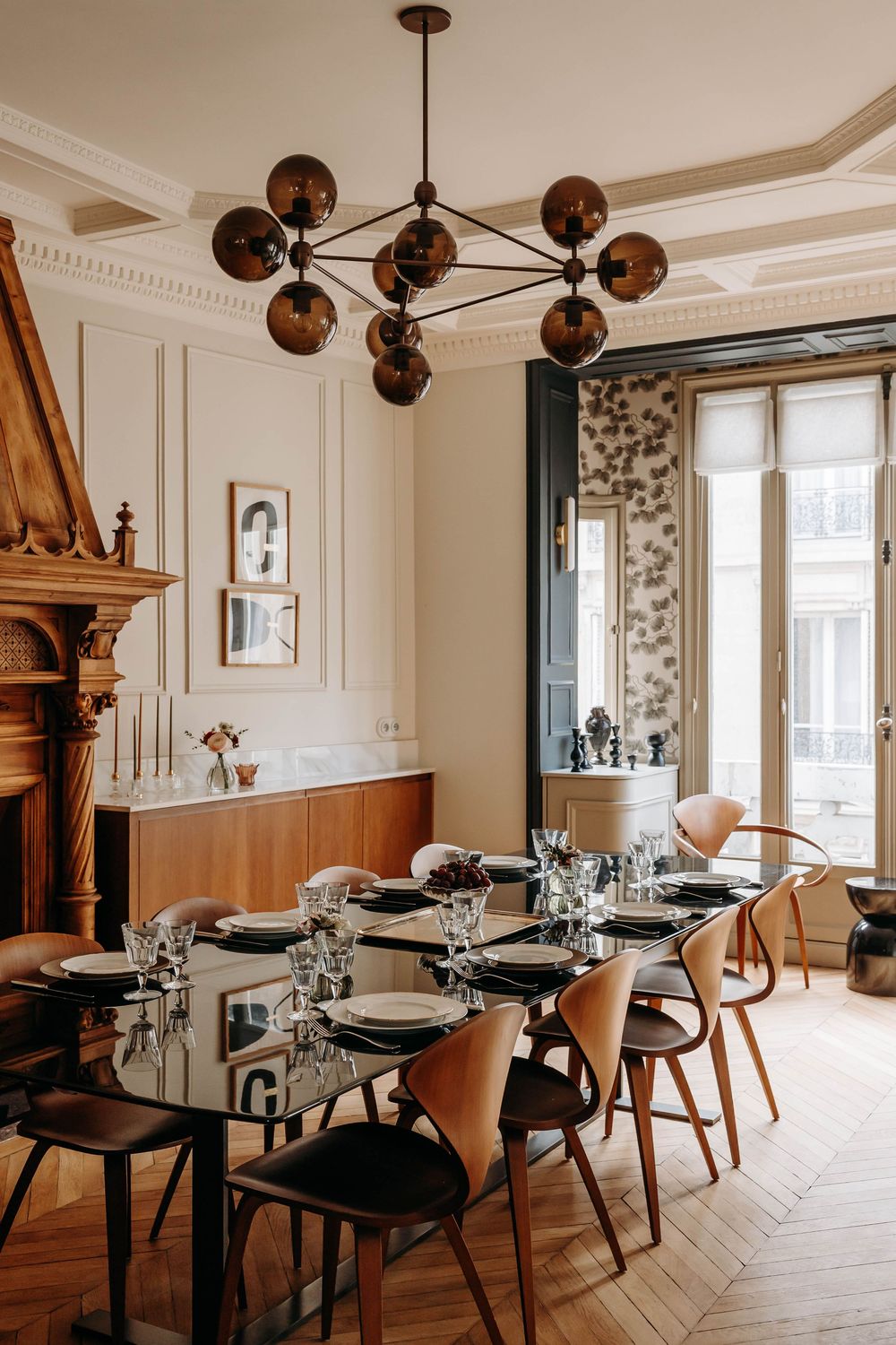 Elegant dining room with glass table, leather chairs, ornate fireplace, herringbone floors, and a modern globe chandelier.