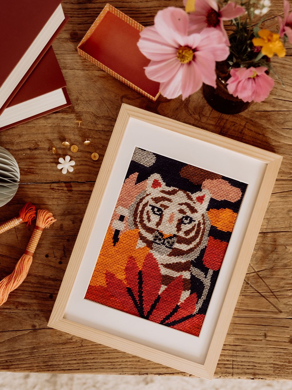 Framed tiger cross-stitch needlepoint art in warm autumn tones displayed on a wooden table with flowers and thread.