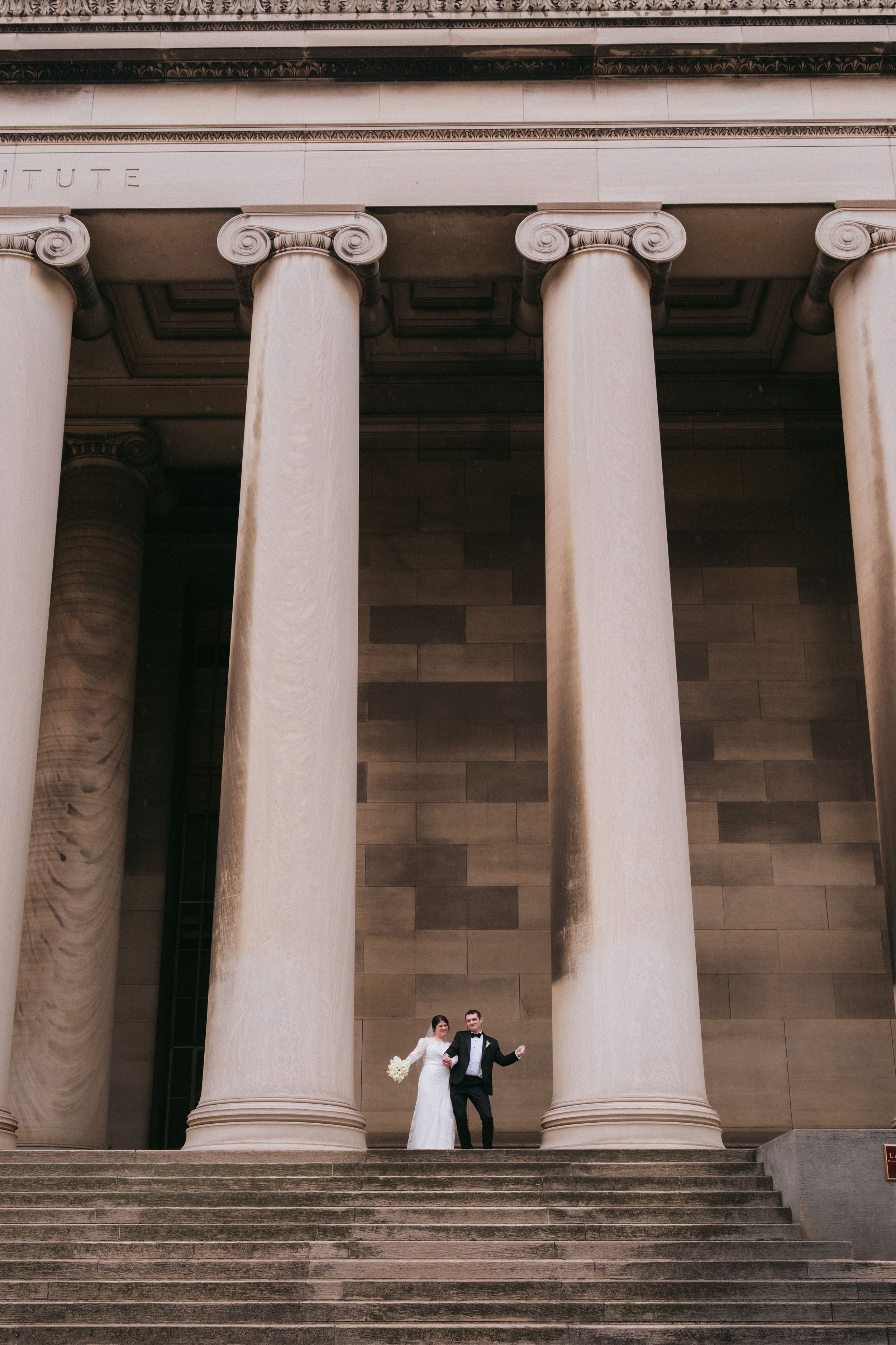 Elegant wedding couple stands between grand white columns on stone steps of historic building facade.