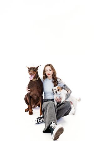 A woman sits with two dogs - a brown doberman and a white small breed - against a white background.