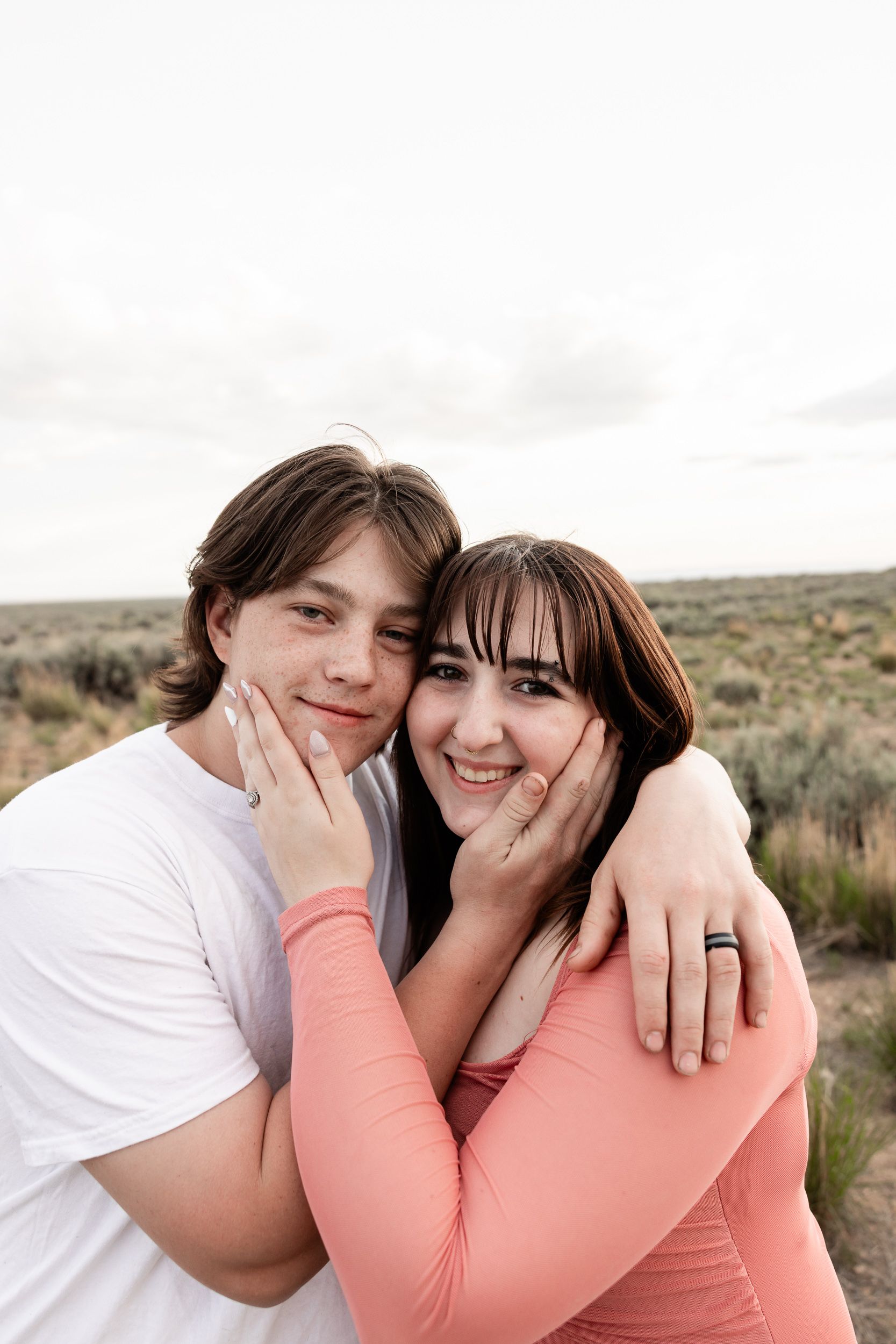 A romantic couple embraces in an outdoor setting with desert brush and scrubland in the background.