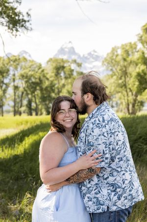 Happy couple embraces outdoors in a sunny field with green trees in the background during a summer photoshoot.