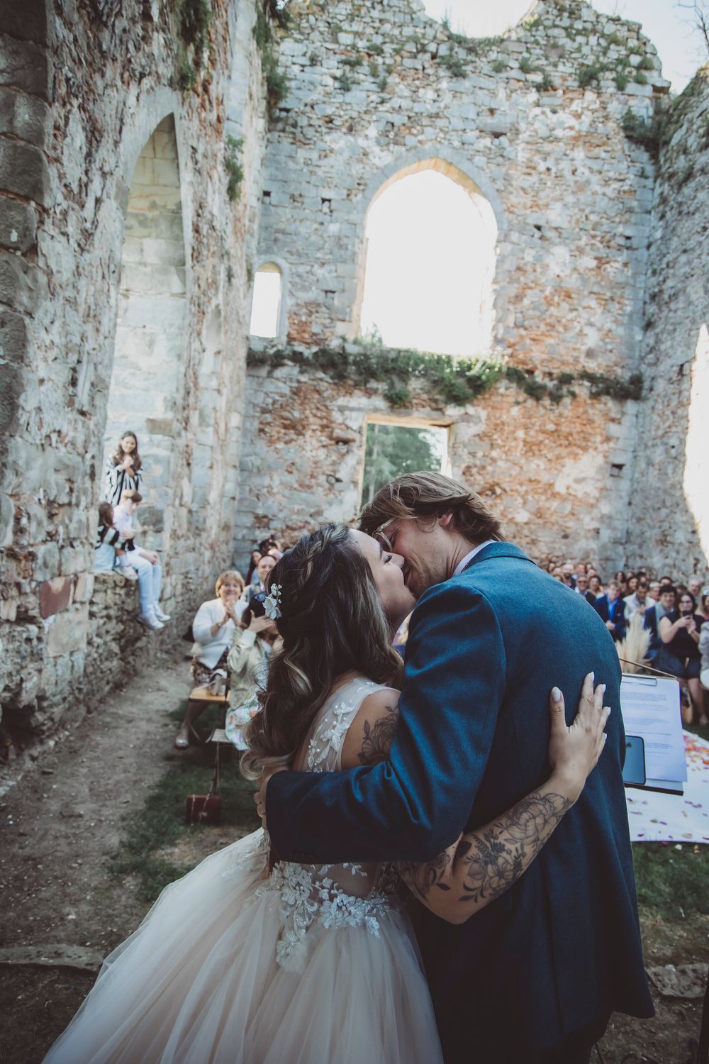 A romantic wedding ceremony kiss in the ruins of an old stone church with exposed brick walls and gathered guests.