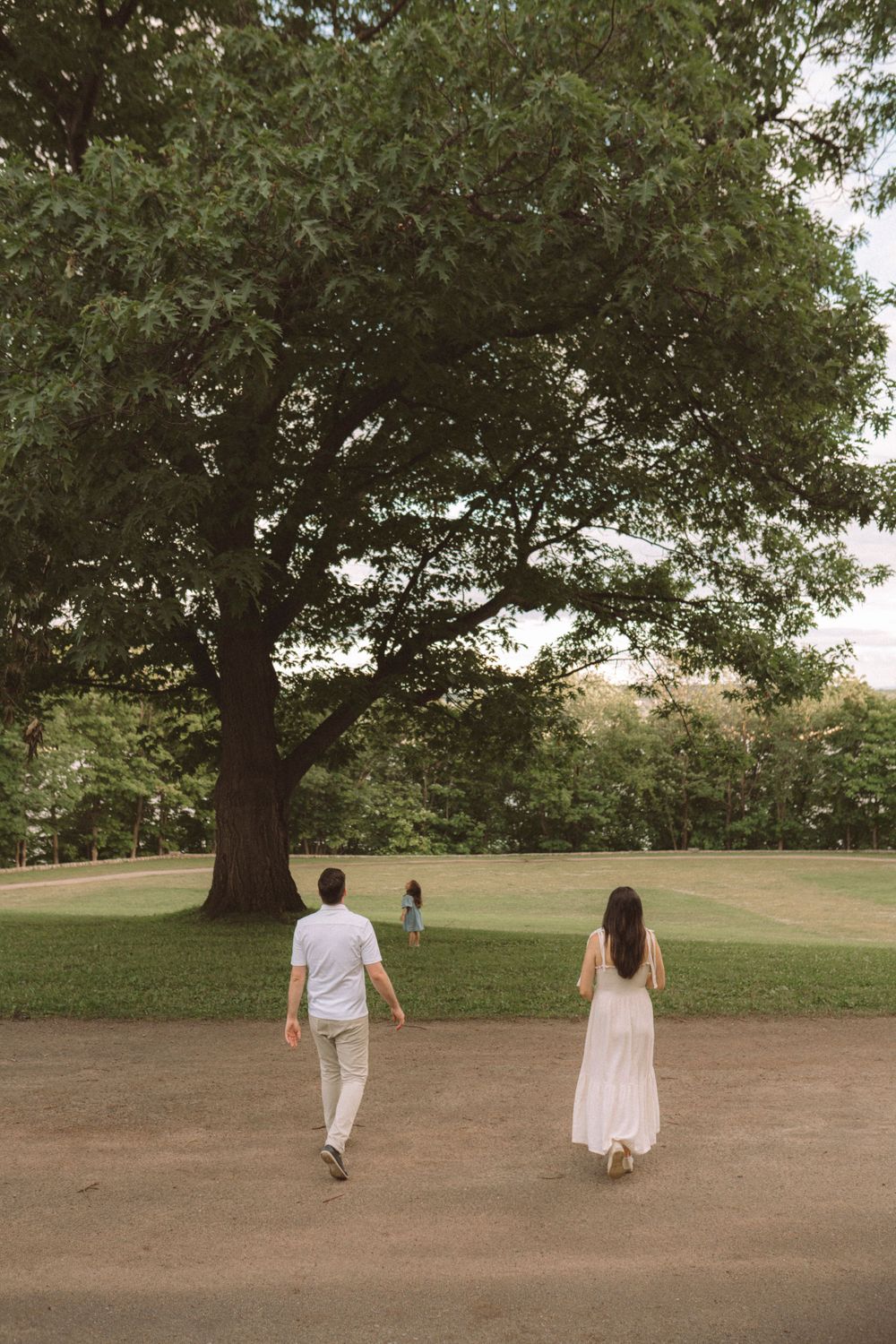 Two people in light colored outfits walk along a tree-lined path on a summer day.