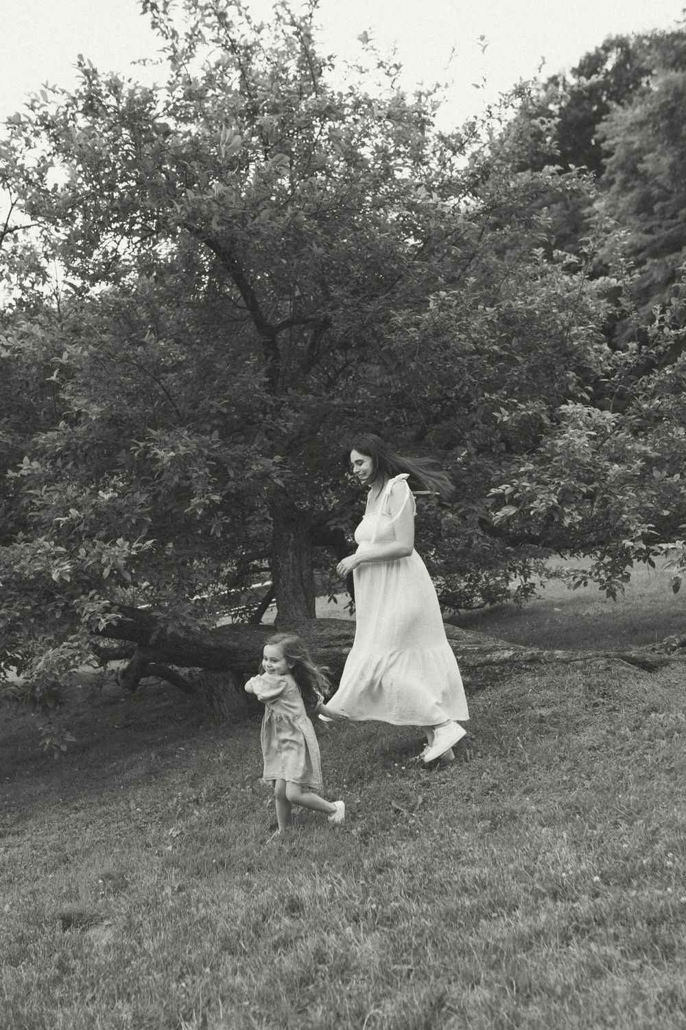 Black and white image of two people in flowing dresses dancing in a grassy field beneath a large tree.