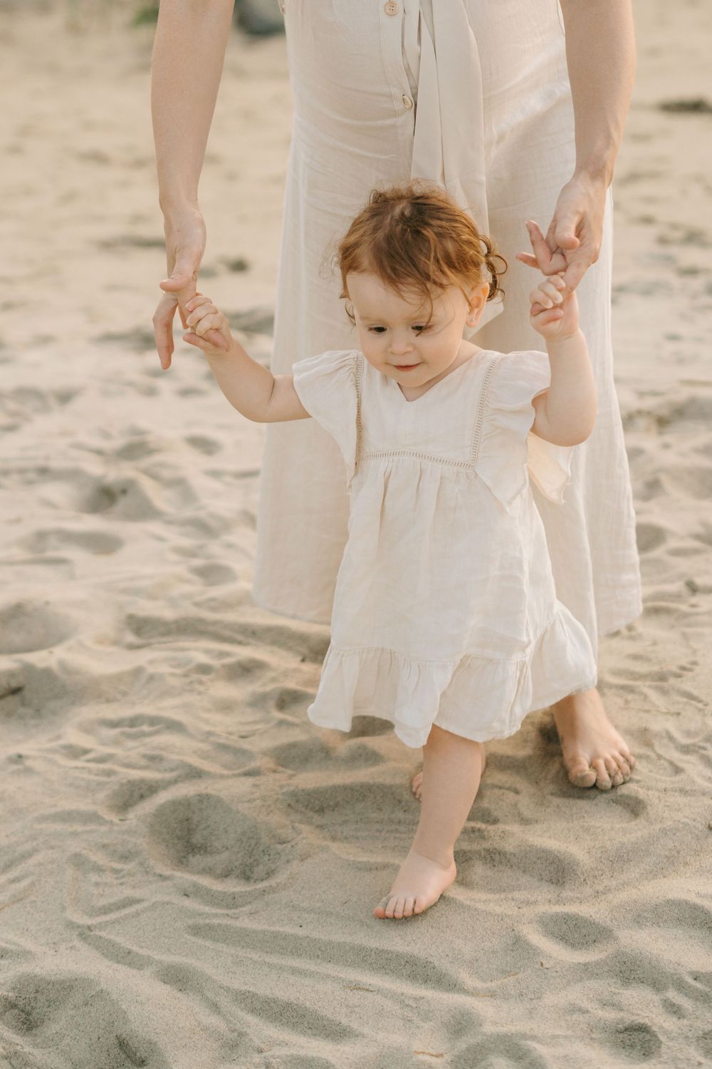 Young child in white dress walking on beach sand while holding hands.