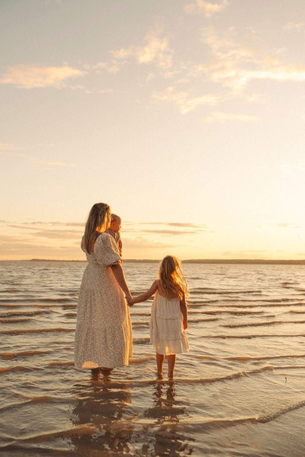 Two figures in white dresses stand together at sunset in shallow ocean water creating a serene beachside moment.