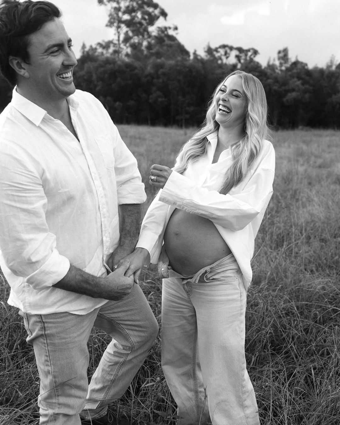 A couple in white clothing shares joyful candid moments together in a grassy field in black and white photography.