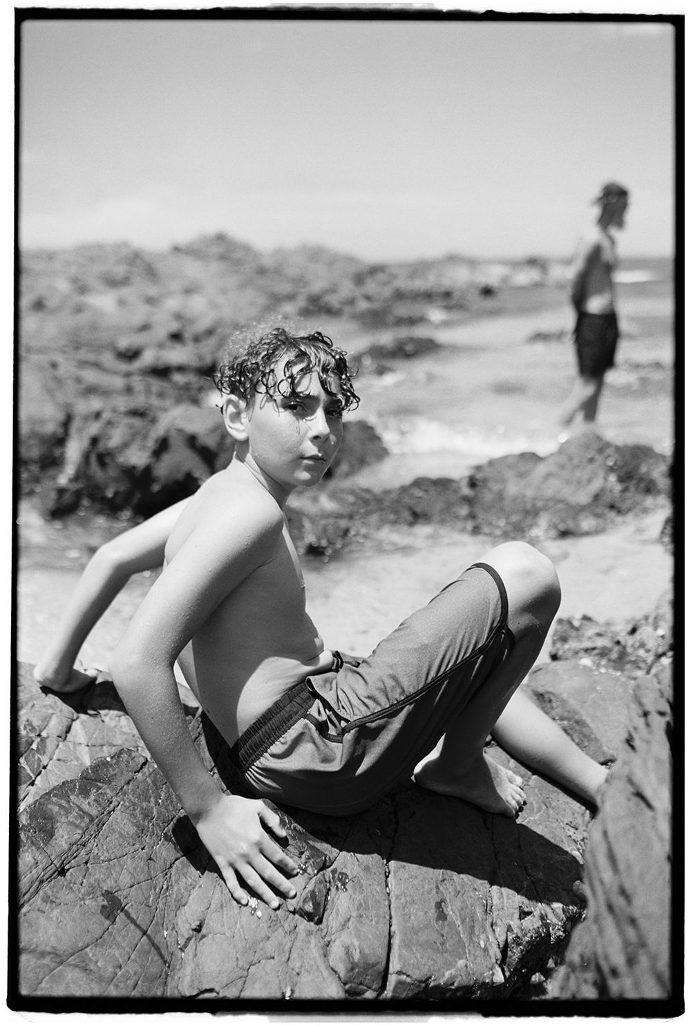 A shirtless boy sits on rocky beach in black and white photo, with another person standing on rocks in background.