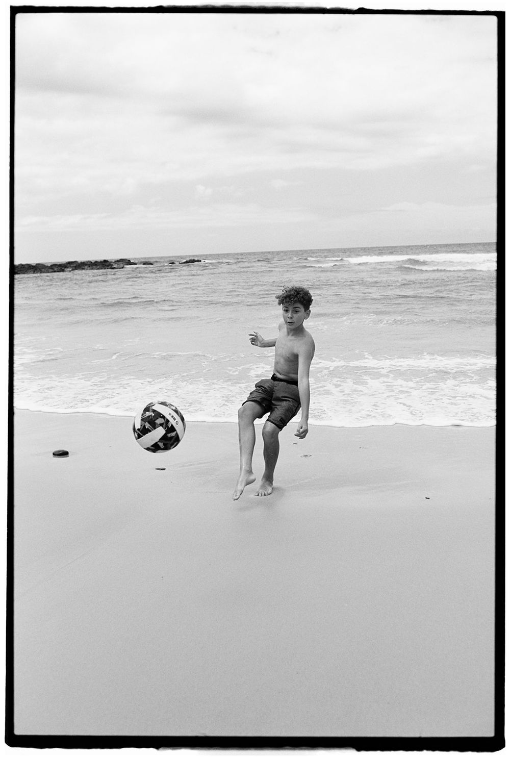 Black and white photo of a shirtless boy kicking a soccer ball on a sandy beach with ocean waves behind him.